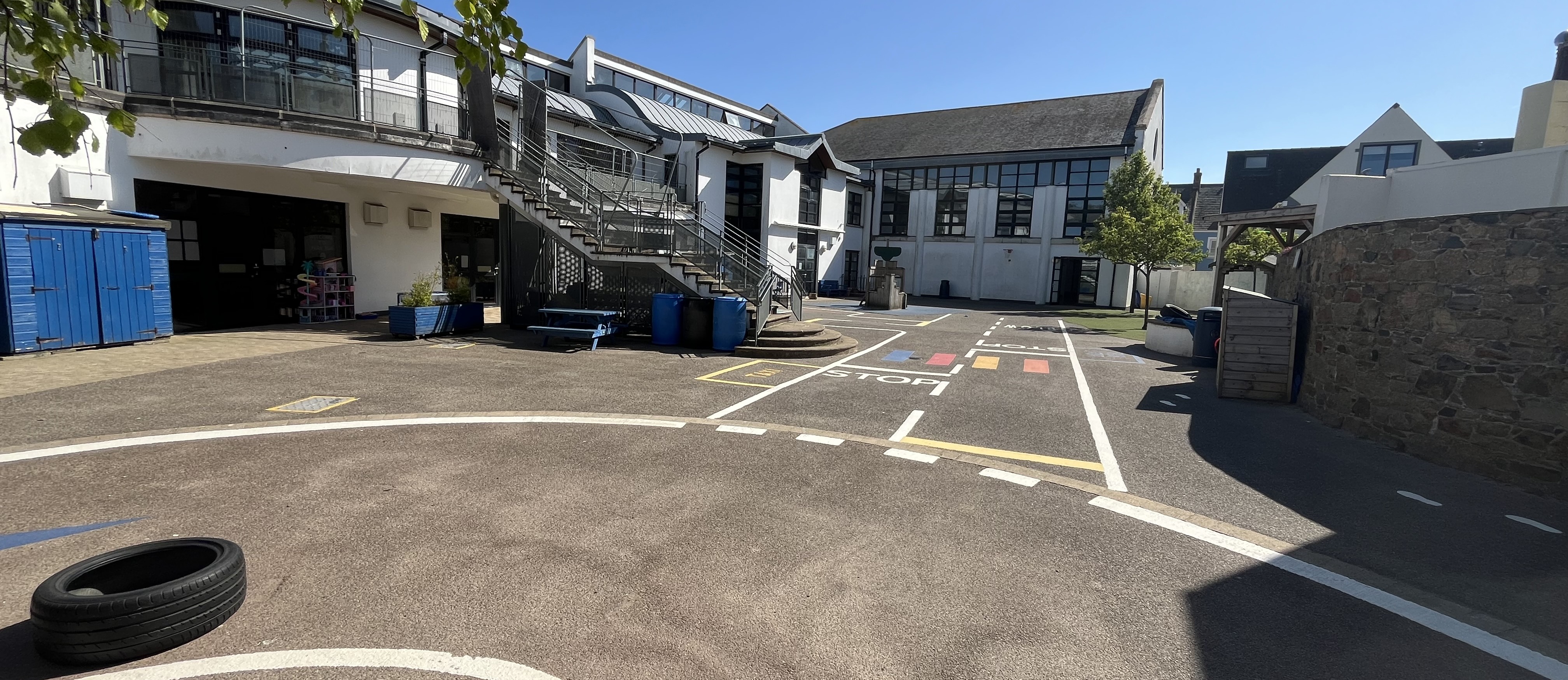 School courtyard with painted road markings and a staircase leading to upper-level classrooms, surrounded by white buildings under a clear sky.