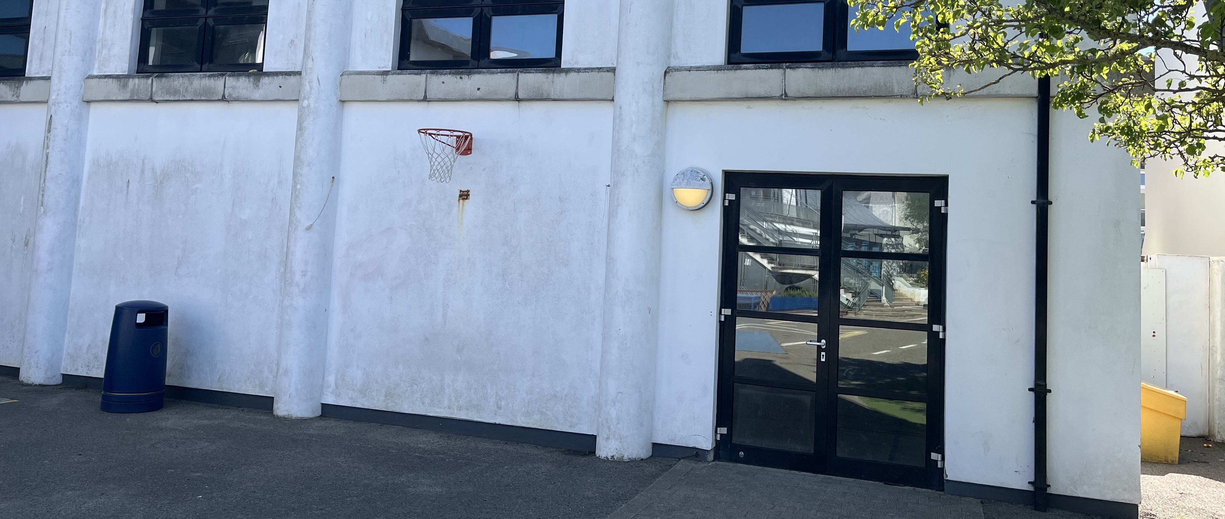 Exterior view of a school building wall with a glass double door entrance, a wall-mounted light, and a basketball hoop above on a paved area.