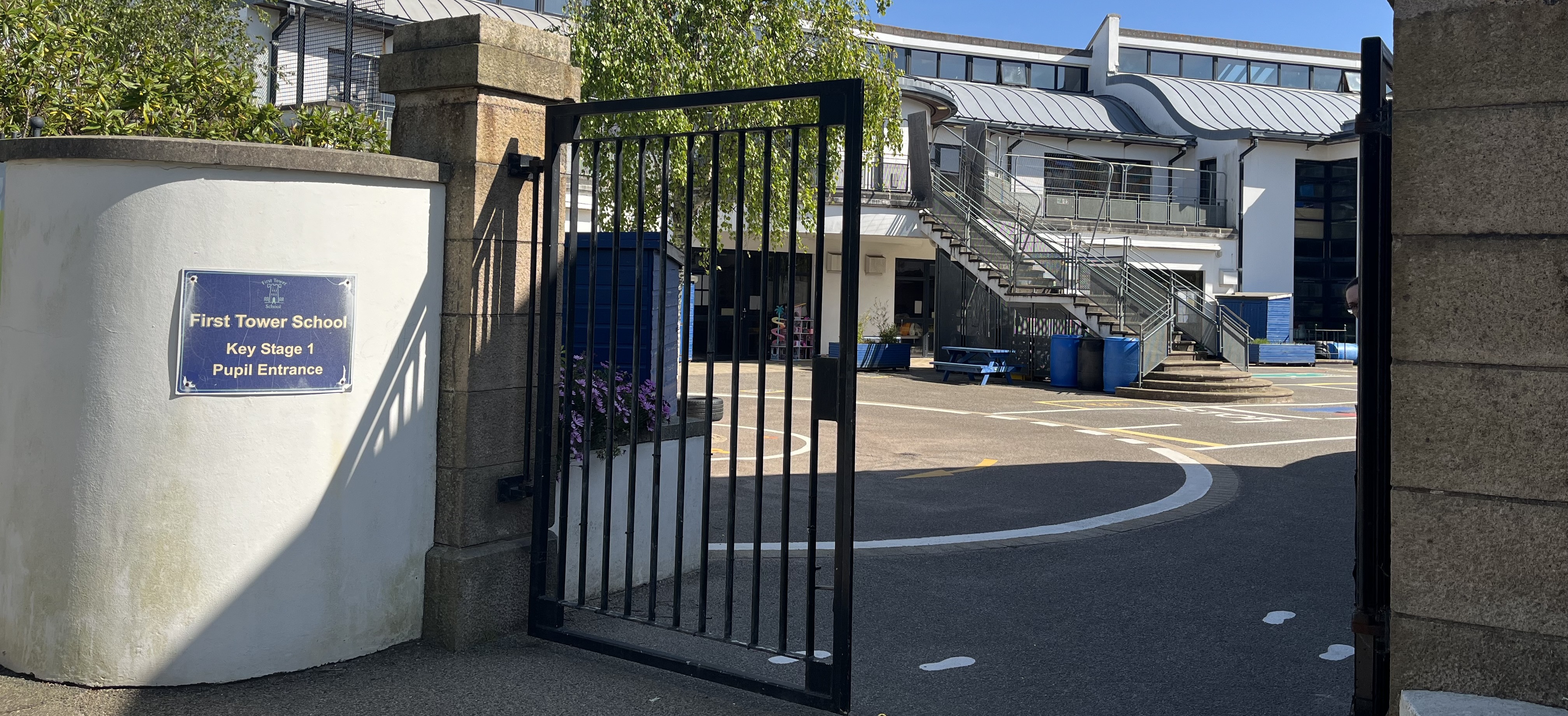 Open metal gate leading into a school courtyard with painted ground markings, stairs to upper levels, and a sign reading “First Tower School Key Stage 1 Pupil Entrance.”
