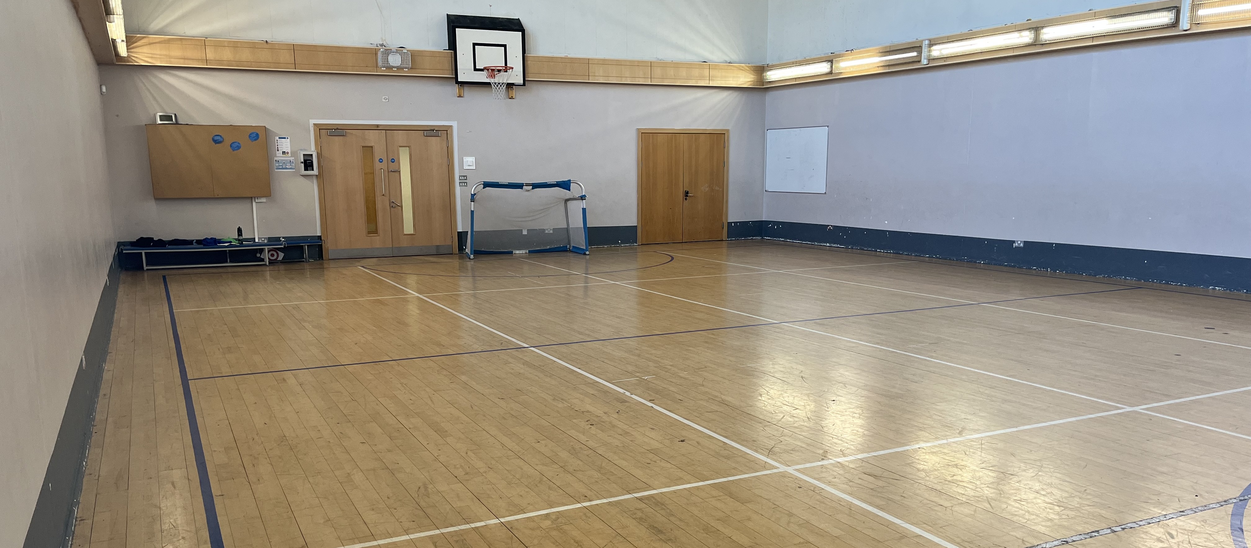 Indoor sports hall with wooden floor markings, a basketball hoop, a small goal, and doors along the far wall under bright lighting.