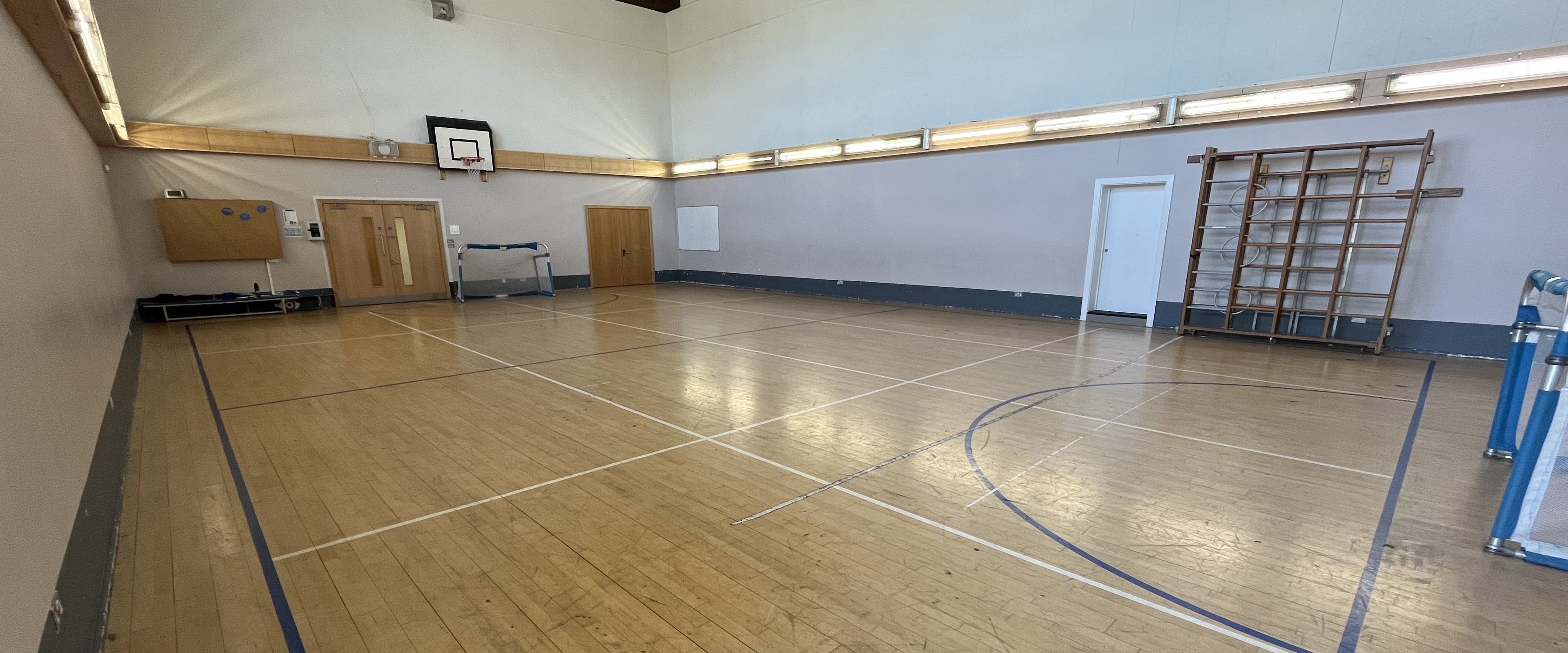 Indoor sports hall with wooden floor markings, a basketball hoop, a small goal, and doors along the far wall under bright lighting.