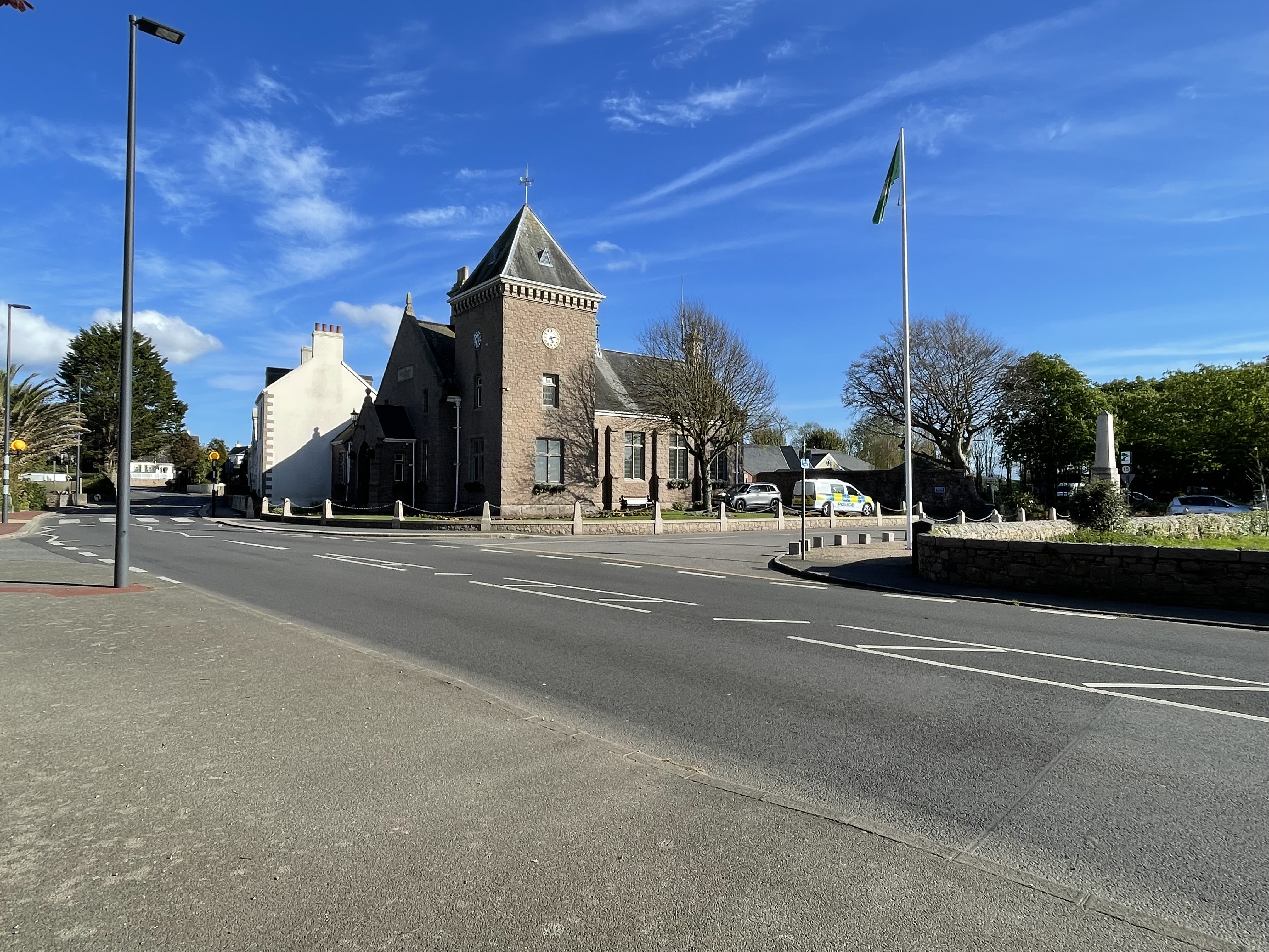 Stone parish hall with a clock tower at a road junction, with a flagpole, trees and parked vehicles under a clear blue sky.