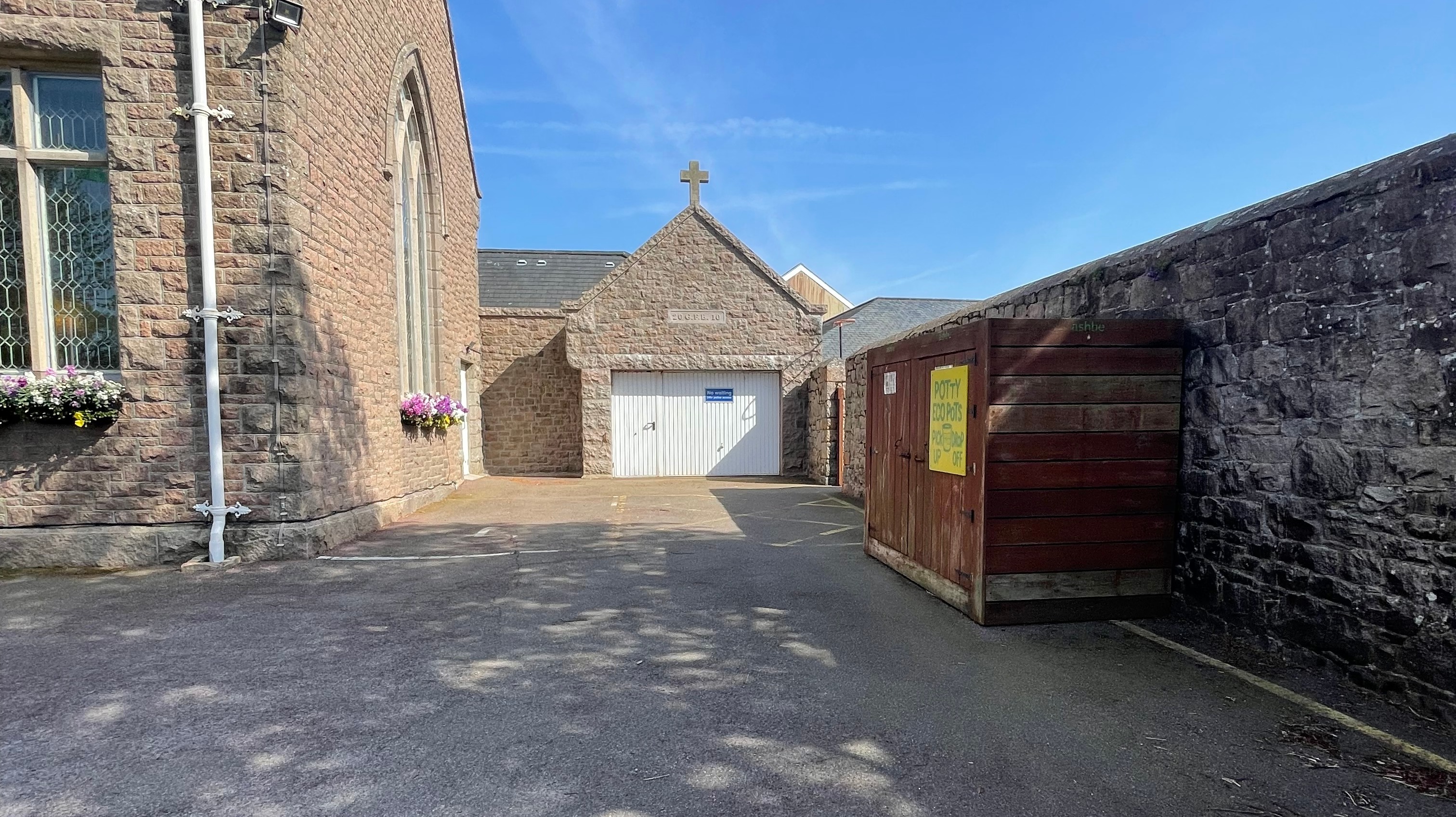 Stone building courtyard with a marked parking area, garage-style entrance door, and a wooden bin store beside a stone wall under a clear sky.