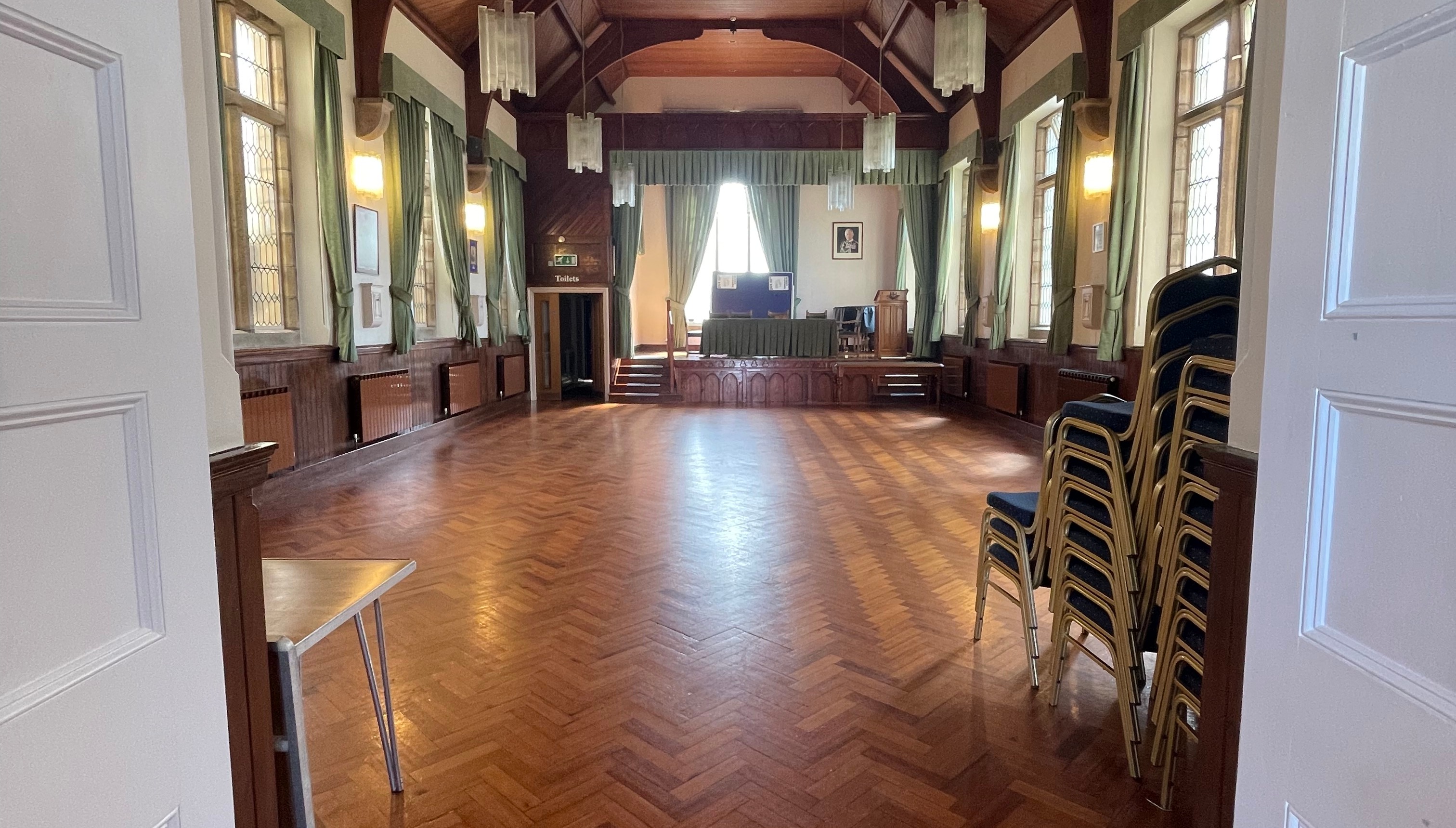 View into a parish hall with polished wooden floor, high ceiling, and a raised stage at the far end; stacked chairs are positioned to one side.