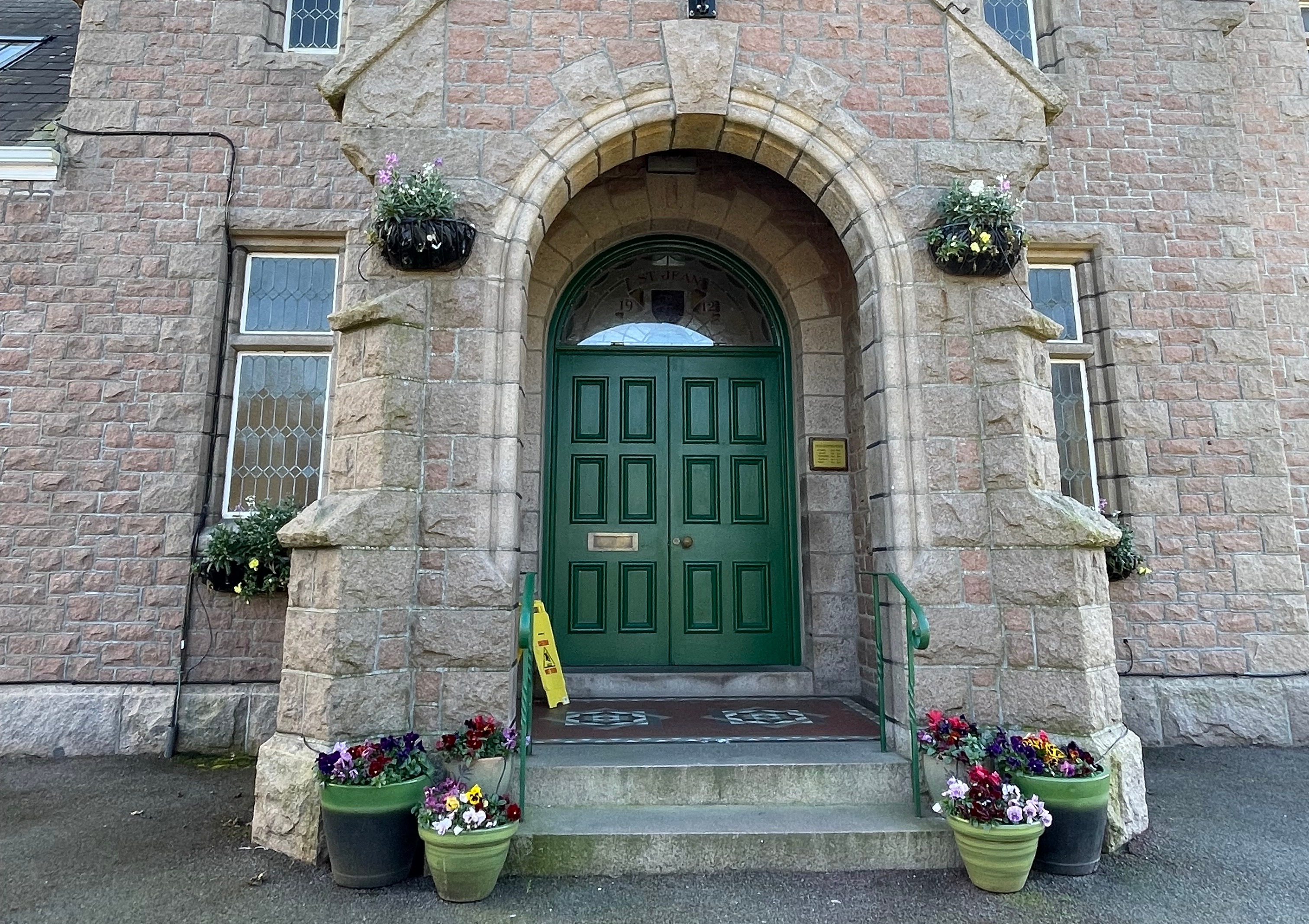 Stone building entrance with a green double door set within an arched doorway, with steps, railings, and potted flowers on either side.