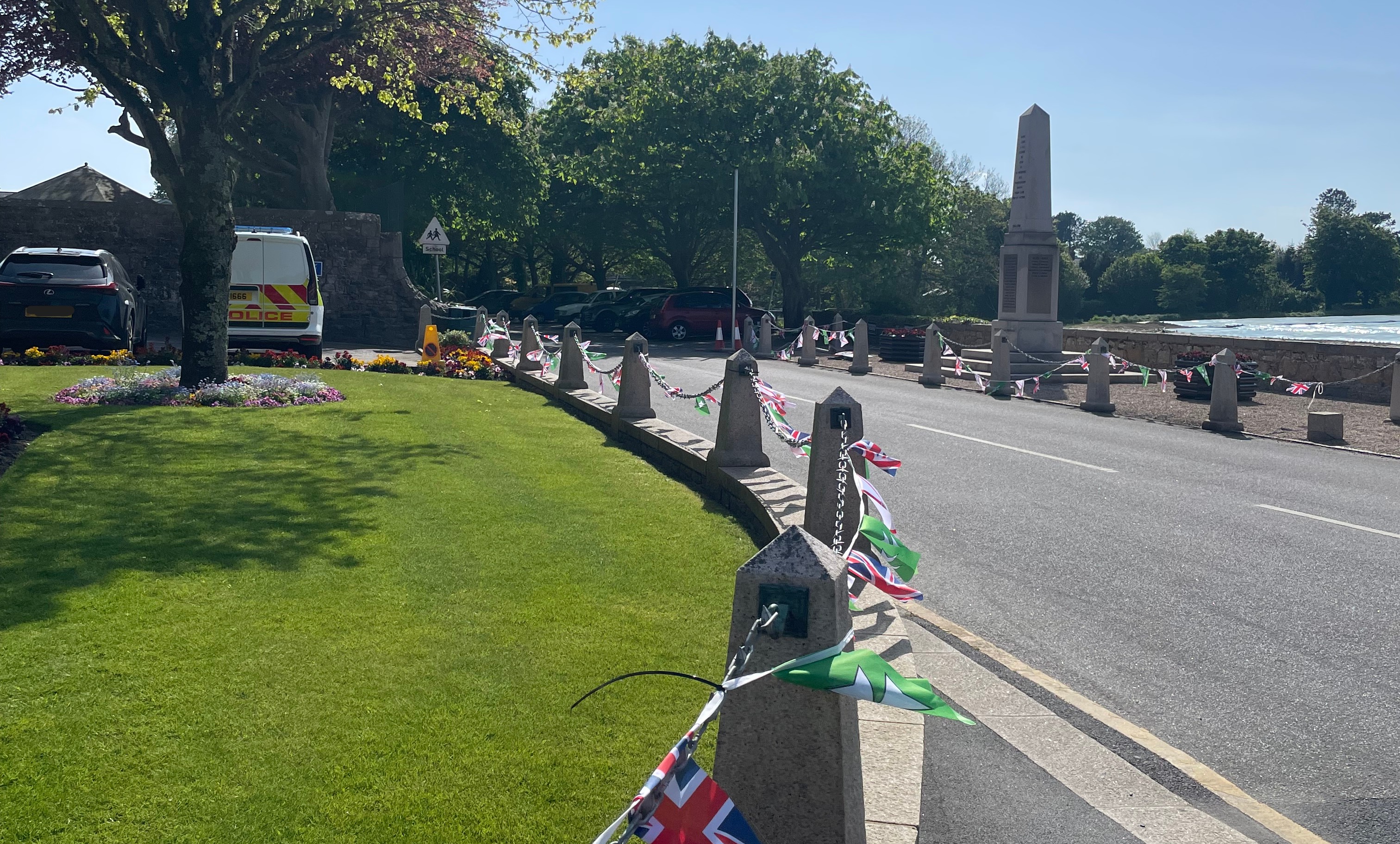 Roadside view with a grass verge, stone posts decorated with bunting, a war memorial, and parked vehicles near trees on a sunny day.