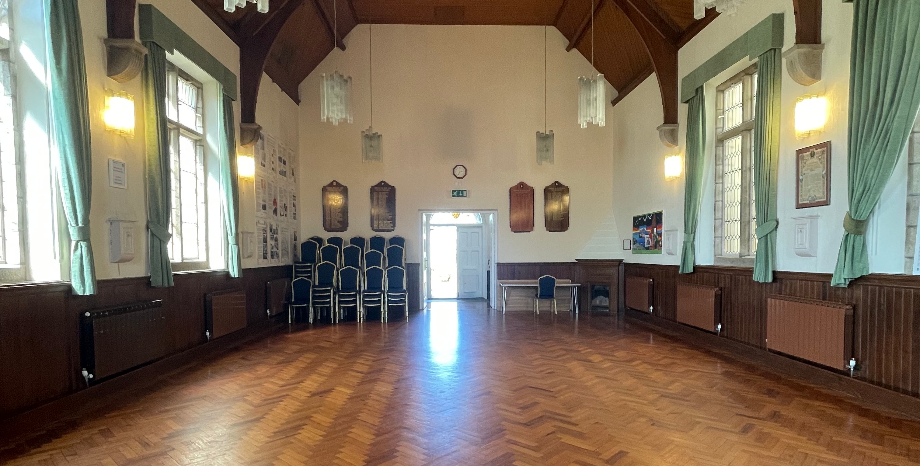 Large hall interior with wooden flooring, tall windows with green curtains, notice boards on the wall, and a stack of chairs near an open doorway.