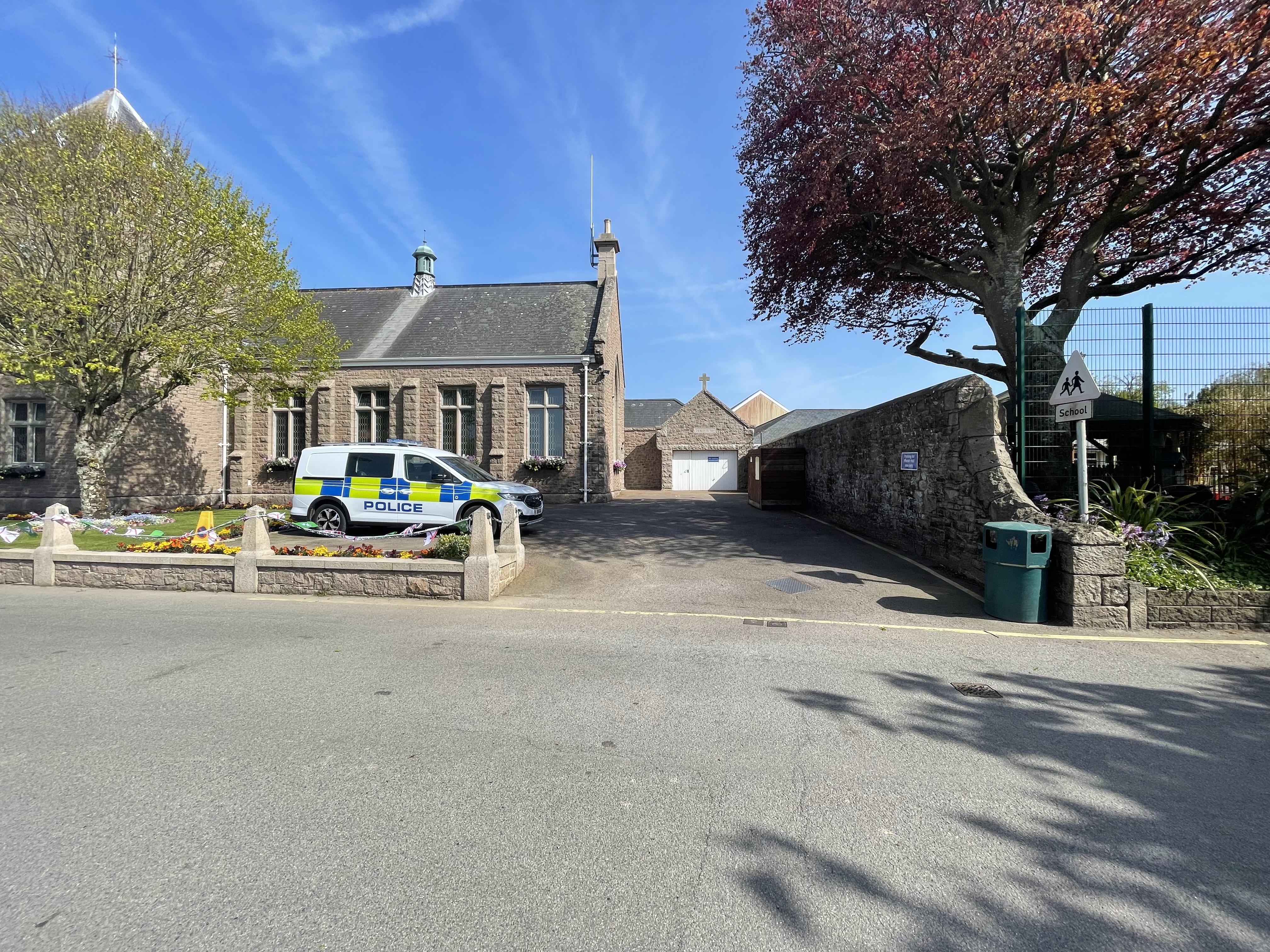 View of a stone parish hall and entrance area from the road, with a police vehicle parked nearby and a driveway leading to a courtyard.