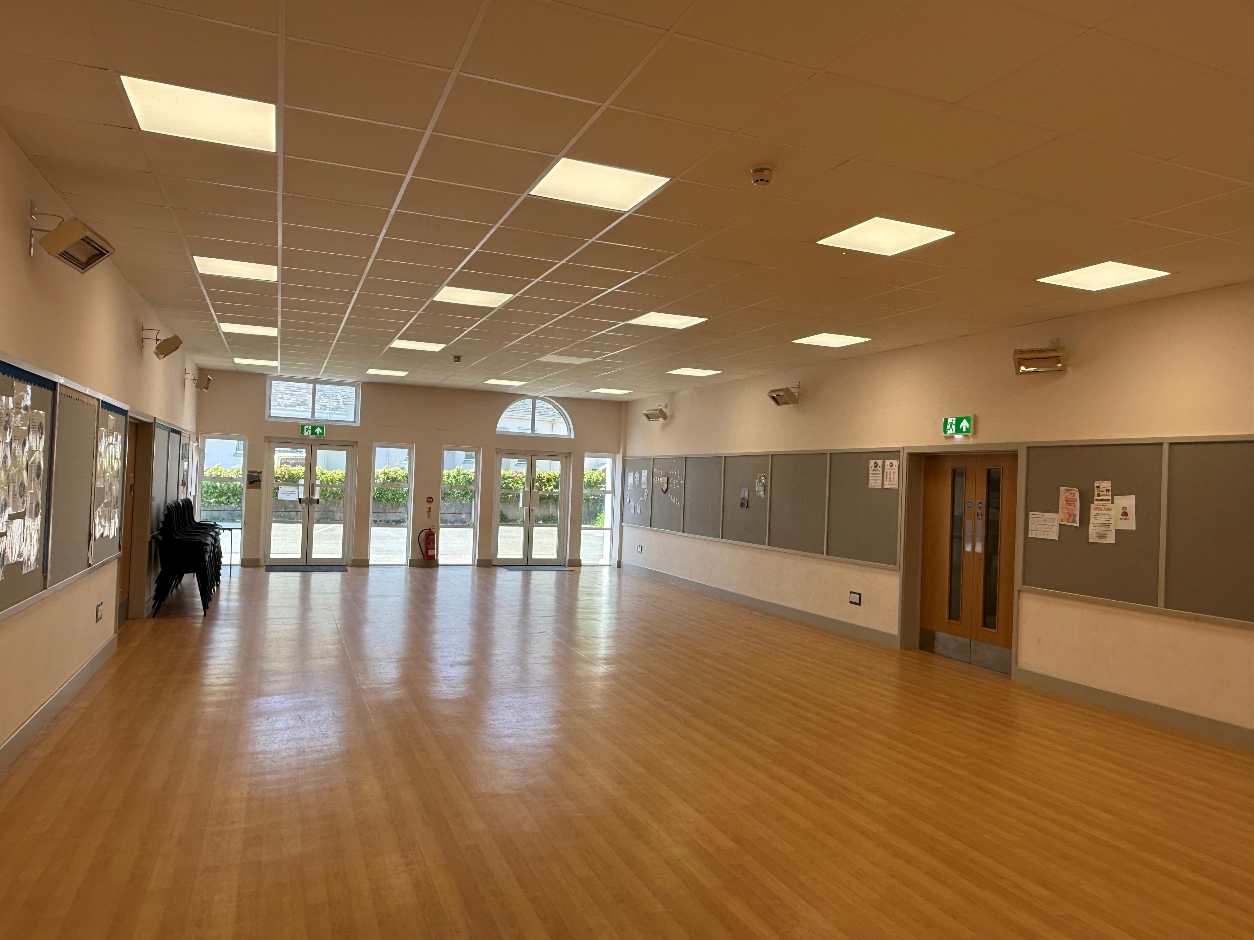 Interior of the Youth Centre Hall, with a wide open floor, bright ceiling lights, and wooden flooring. Double glass exit doors are visible at the far end, with emergency exit signs above.